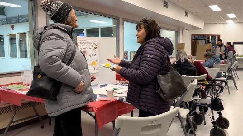 Two women stand talking in a room of tables with display boards and people in chairs.