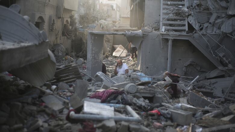 A bearded man wearing a white T-shirt speaks on a phone surrounded by rubble.