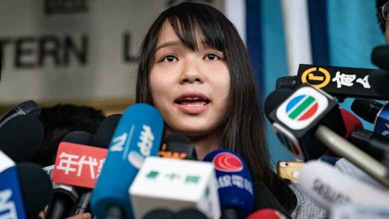 A woman speaks before a large set of microphones.