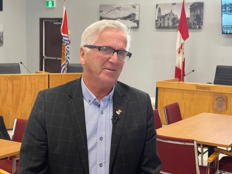 A Caucasian man with white hair, glasses, and a dark striped blazer stands in a hall with Canadian flags behind him. 