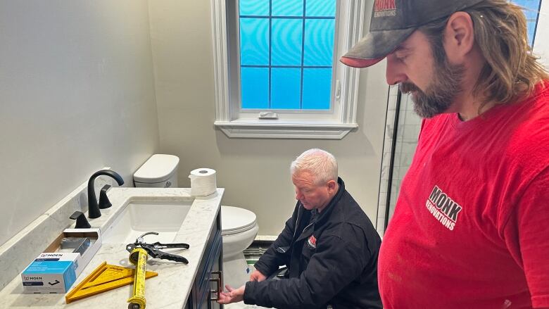 A man in a red t-shirt works on a bathroom renovation next to a man in a black jacket.