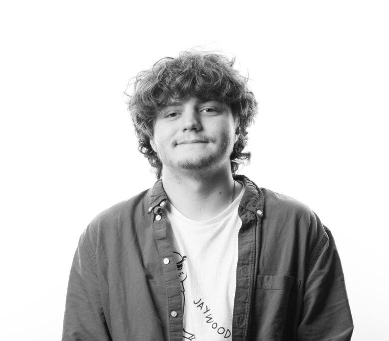 Young man with curly dark hair sits on a stool with his hands clasped at his lap. He wears a button down shirt open with a white graphic T-shirt underneath. He is wearing dress pants.