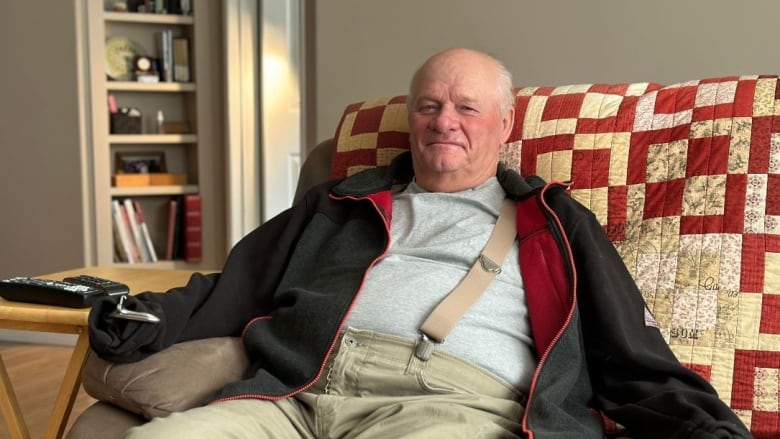 Man in overalls and black sweater with prosthetic hooks sits on couch.