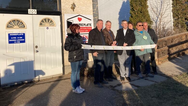 A group of people stand proudly next to a new emergency shelter in Meadow Lake, Sask. They are holding a ribbon containing the Government of Saskatchewan logo.