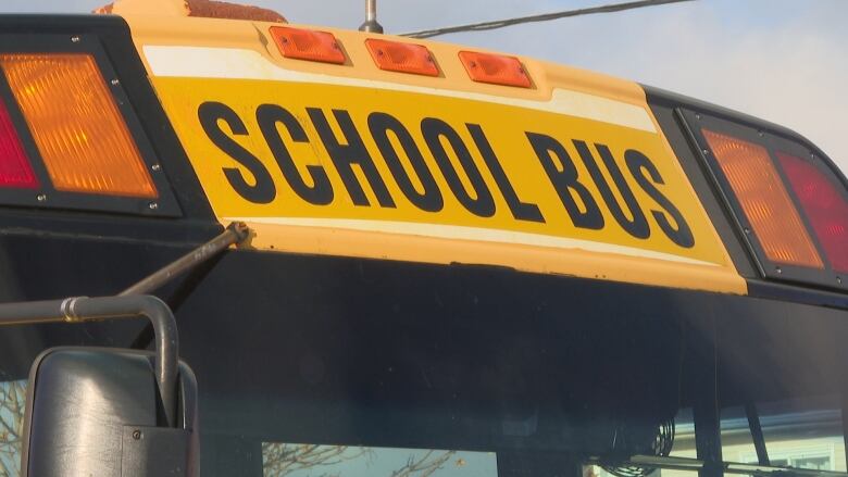 Only the top of a yellow school bus is pictured. It has the words School Bus printed at the front of it. Orange and red lights flank the words. 