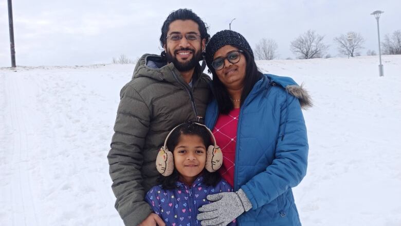 Father, mother, and daughter standing together outside near a snow hill smiling for the camera.