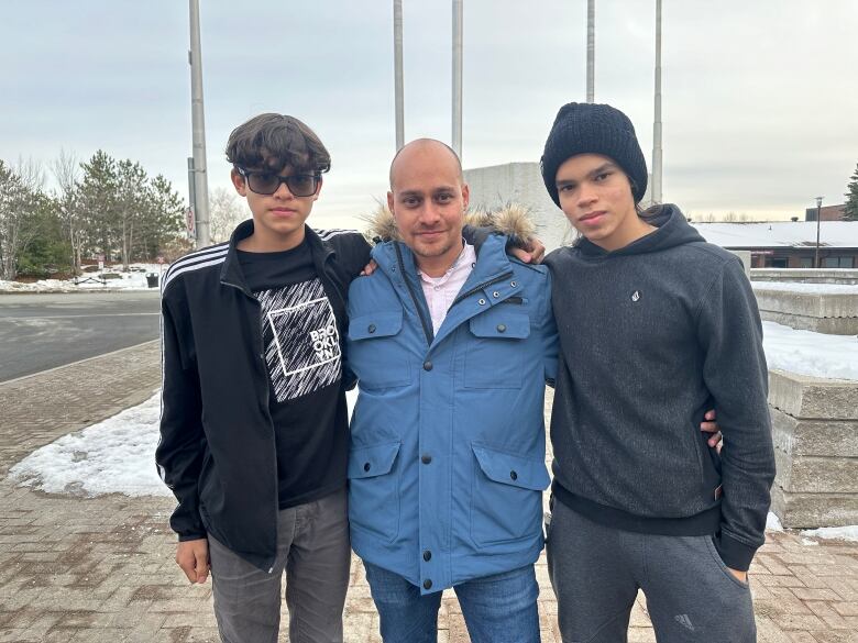 A father and his two sons standing together in a photo in front of college sign. 