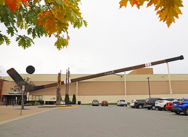 A huge hockey stick lies horizontally next to an arena, behind a totem pole and a row of cars.