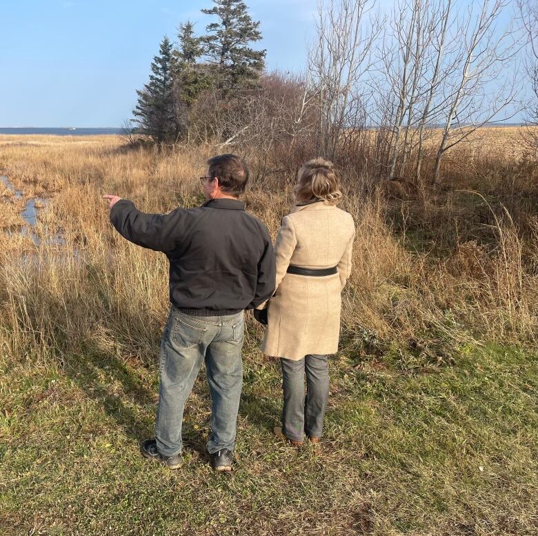 A man and woman stand with their backs to the camera and the man points out to the water 