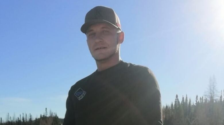 A man is wearing a baseball hat. He is standing outside. His hands are clasped together. 