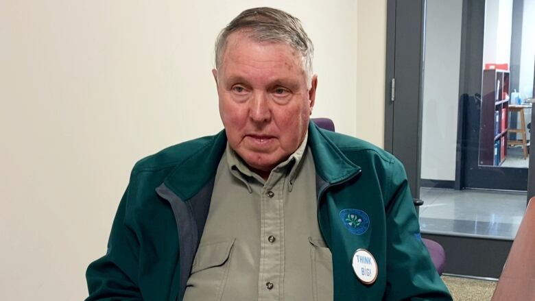 A white man with grey hair and a green jacket sits in a chair by a brown table