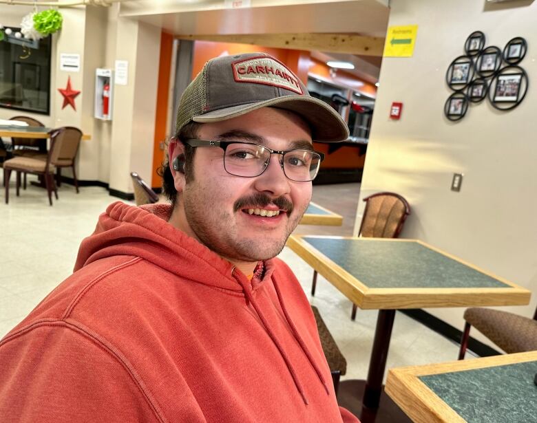 A man in a baseball hat and red hoodie sits in a cafeteria.