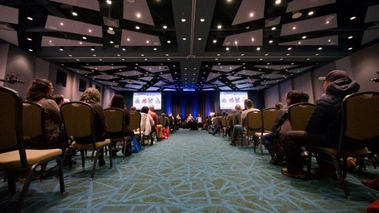 People sit on cushioned chairs in a carpeted conference hall, while about three presenters sit on a stage at the front of the room