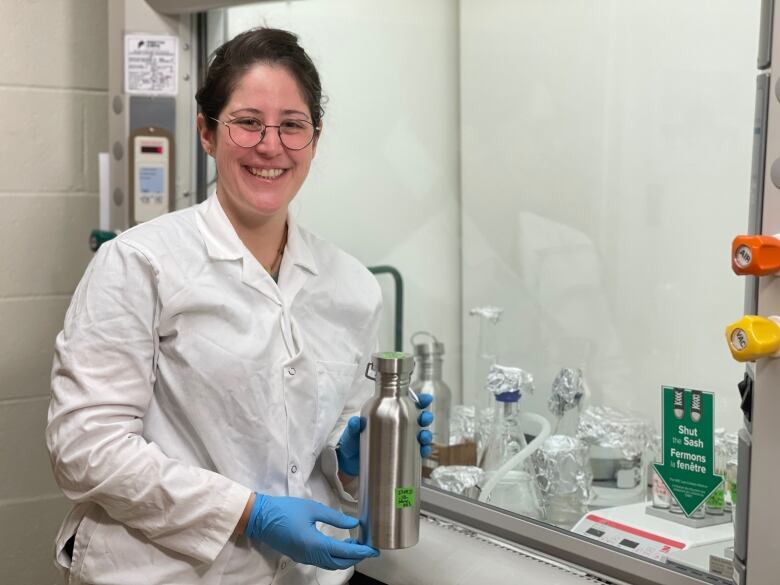 A young woman wearing a white lab coat and blue gloves holds a silver canister in a science lab.