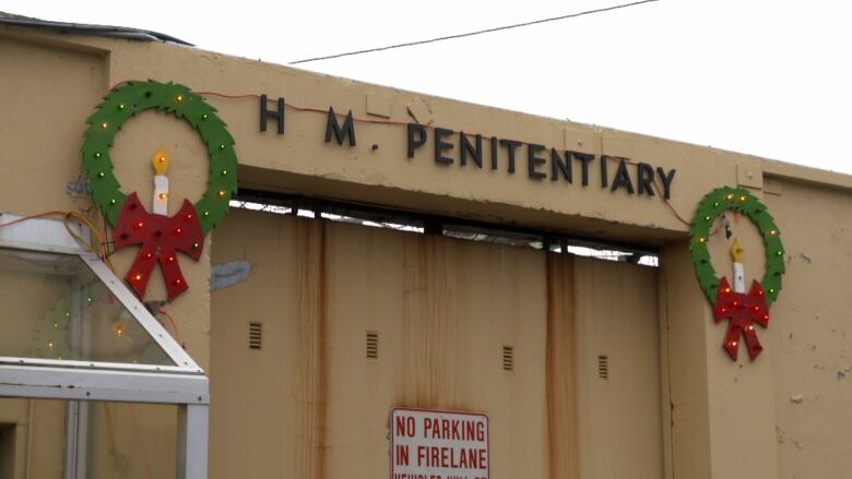 The entrance to a jail with a sign that reads 'H M PENITENTIARY', with two wooden wreaths hanging on either side of the sign.