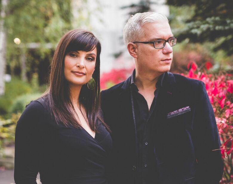 A man and woman in dark clothing stand outside in front of a bush with bright red leaves.