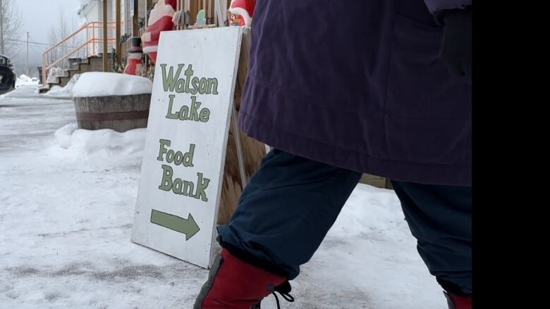 A person's legs are seen walking into a building with a sign, 'Watson lake Food Bank.' 