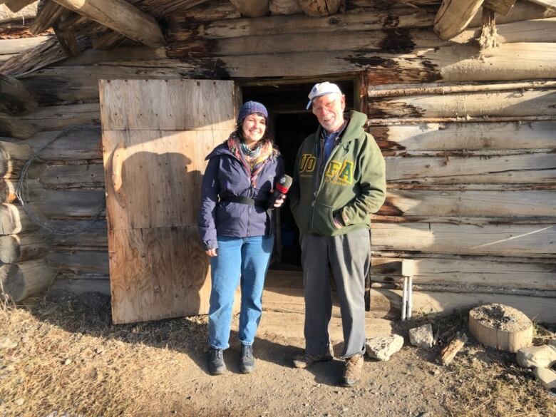 A white woman holds up a mic to a white man standing outside a small shack-like structure.