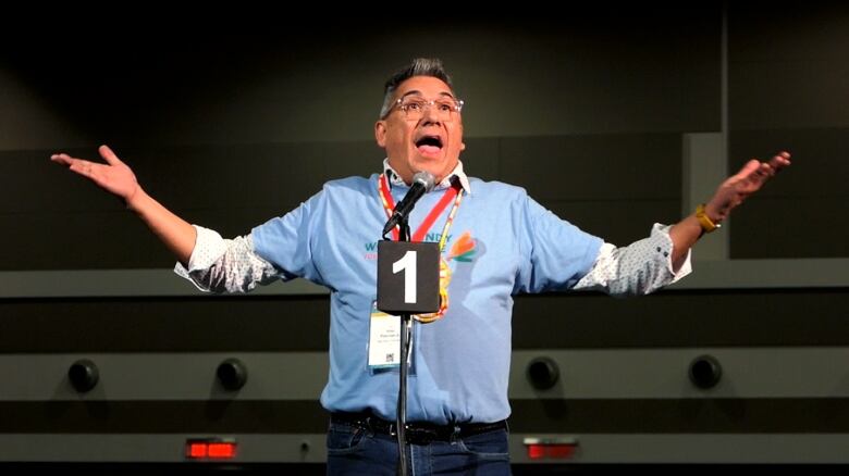 An Indigenous man speaks and gestures into a microphone at an events centre.