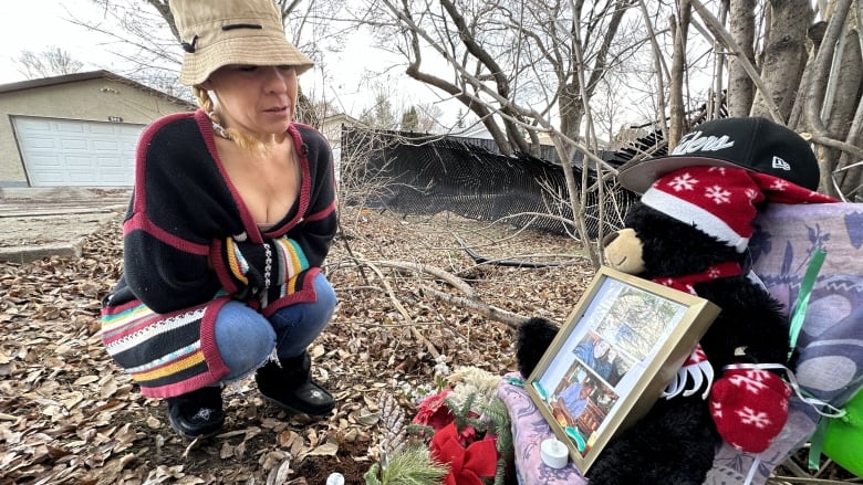 A woman crouches on leaf-strewn ground with a garage in the background. She's looking at an arrangement of items that include a teddy bear, flowers and photos.