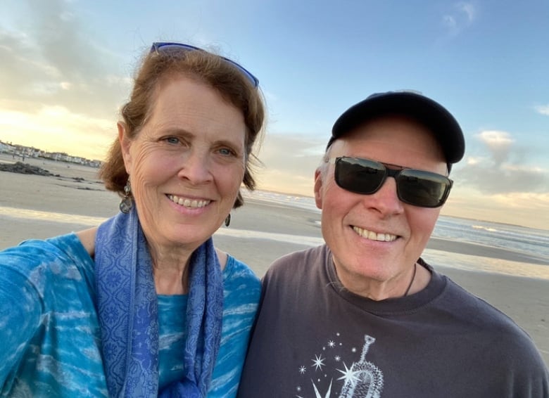 A woman and man smile at the camera with a beach behind them. 