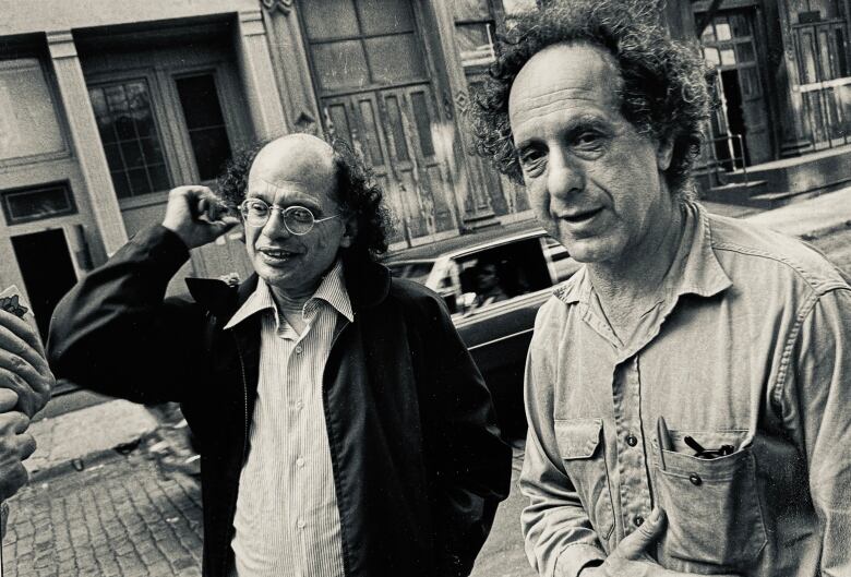 Two men are seen on a New York Street. The one on the right has curly hair, and stares directly into the camera, wearing what appears to be a denim button up shirt. On the left, A man who is balding, but still has long hair on the sides, twists his ear, and looks away from the camera, further down the street. He wears a pinstripe shirt and a jacket. 