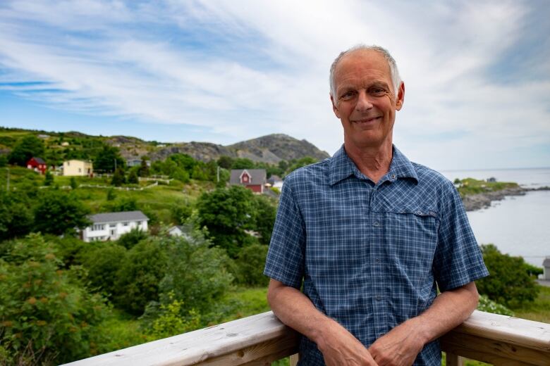 A man in a plaid shirt stands on a porch overlooking the ocean.