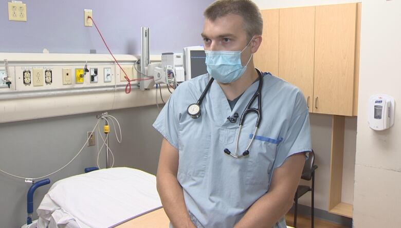 A man wearing scrubs and a surgical mask stands in a hospital room.