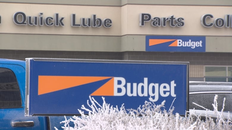 The front of a grey corporate building is in the background with a large cargo van in front of it. The building and the van have the orange and blue Budget logo on them.