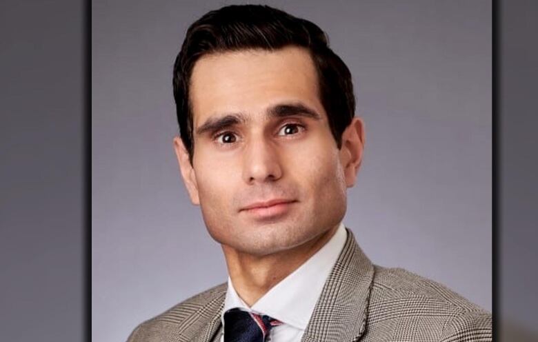 A man with short black hair and a beige blazer looks into the camera.