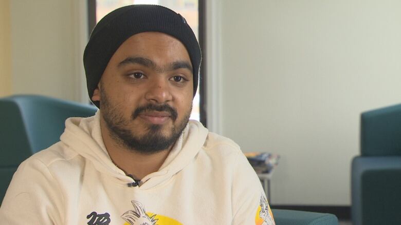 A man with a beard wearing a white hoodie and black beanie sits in a waiting area.