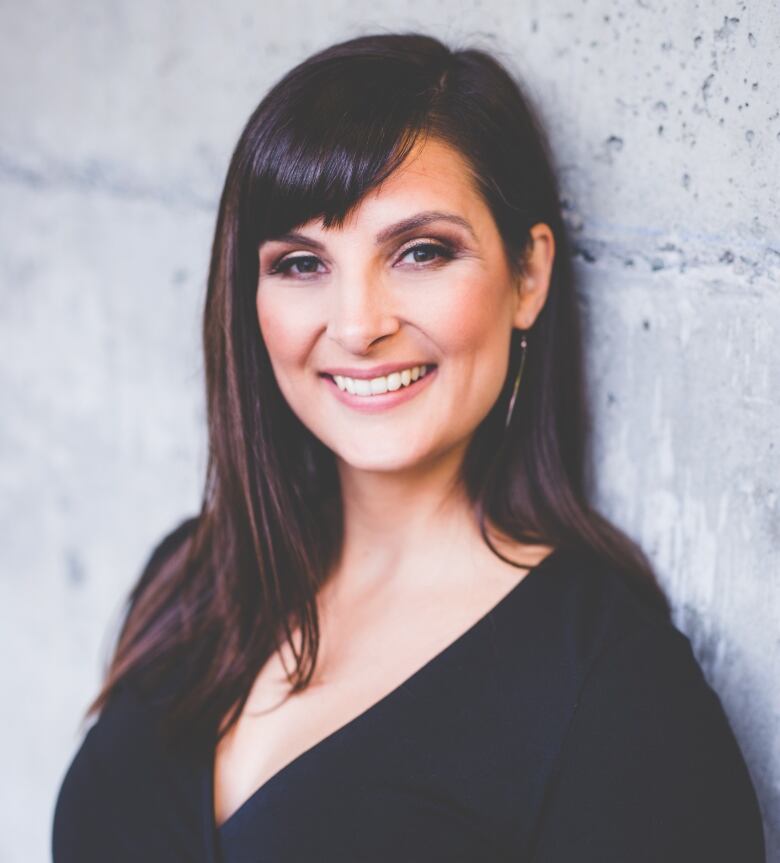 A smiling woman with long dark hair in a dark dress leans against a wall.