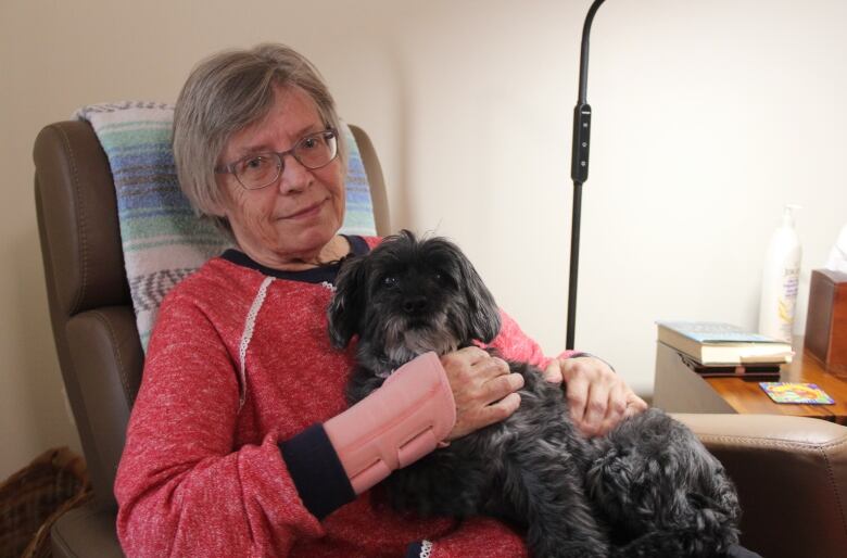 A woman in a red sweater sits on a chair while one of her dogs lay on her lap.