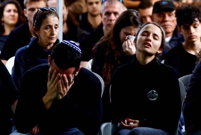 A mourner holds their head in the hands as another closes their eyes and tilts their head up at a funeral.