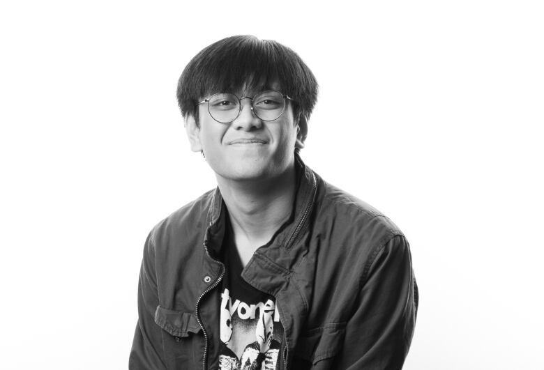 black and white portrait of young man sitting on stool facing the camera. He has short, dark hair with bangs and wears round, wire-framed glasses. He is wearing a dark button-down shirt that is open to a graphic T-shirt underneath. He smiles with his lips closed. 