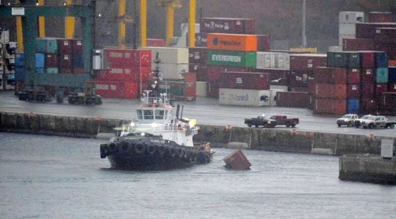 The tugboat with a black hull and white cabin is tied to a floating red shipping container, with the colourful stacks of containers behind it