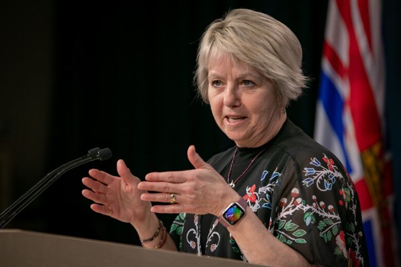 A middle-aged white woman with a blonde bob gesticulates with her hands while standing at a podium speaking. She wears a gold ring on her wedding finger and a gold ring on her right pinky finger. Bracelets adorn her right wrist and a smart watch is on her left wrist. Her top is black with a floral pattern.