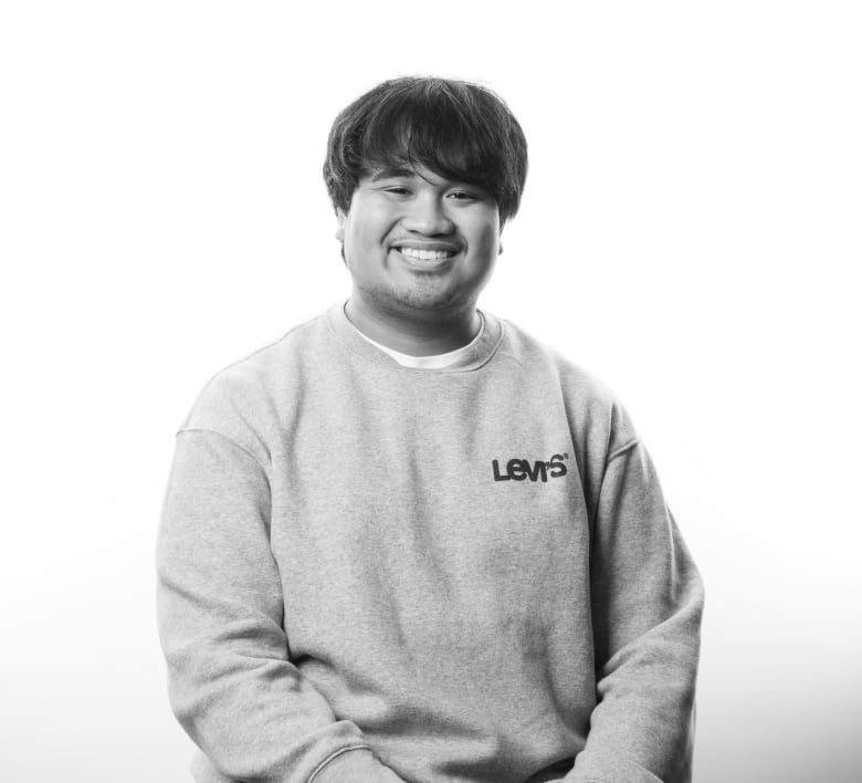 Black and white portrait of young man sitting on stool. He has shaggy, dark hair with bangs and has a broad smile showing his teeth. He is wearing a grey sweatshirt with a Levi's logo on the left chest area. He is wearing dark pants and his hands are resting on his thighs.