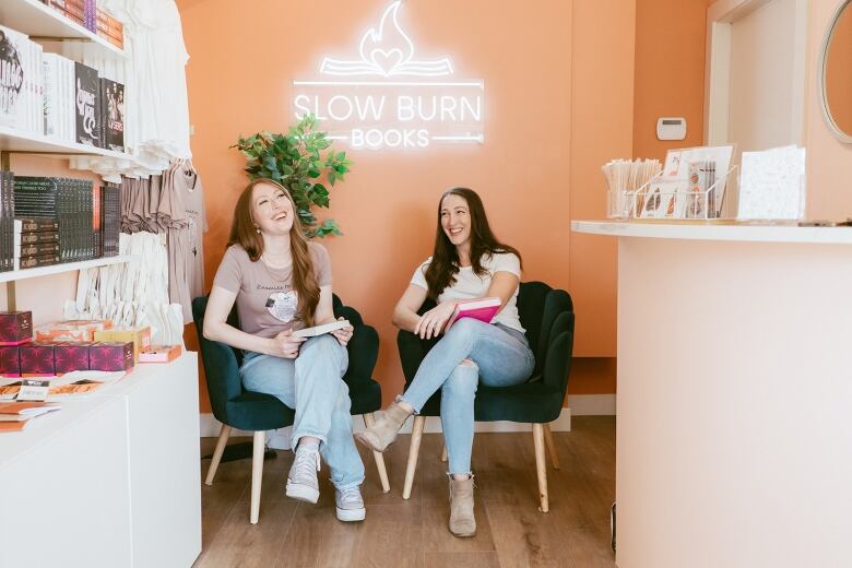 Two women laugh as they sit together in a bookstore with peach-coloured walls and a neon sign that reads 