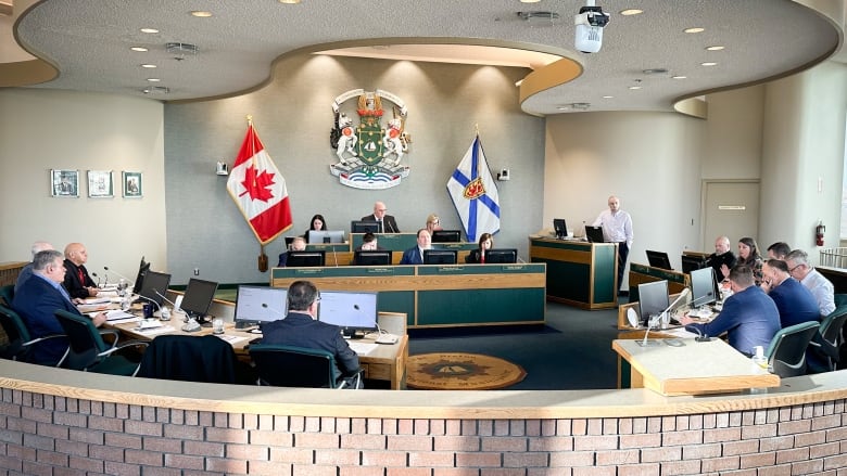 A semi-circular room is shown with a large crest and a Canadian and Nova Scotian flag on the wall and people and computers arrayed around them.