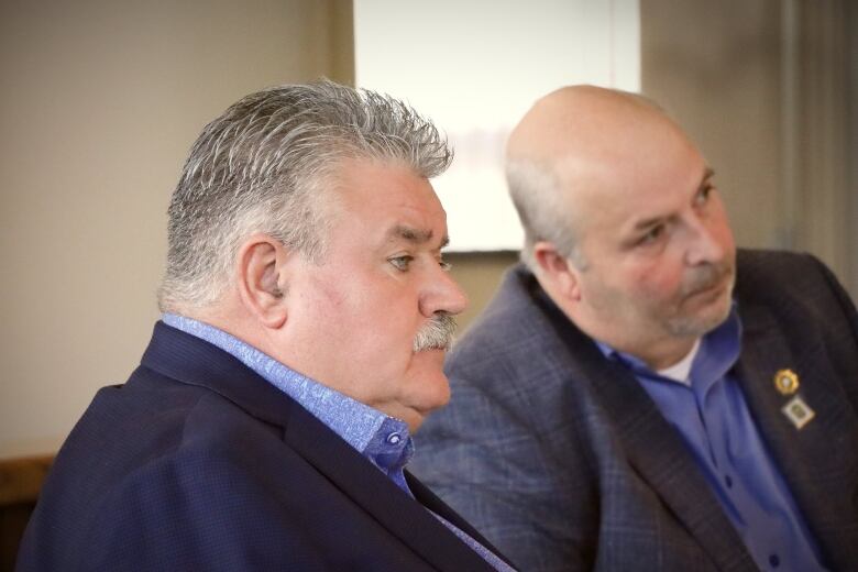 A man with a blue suit and shirt and grey hair and moustache looks away as another man in the background is out of focus.