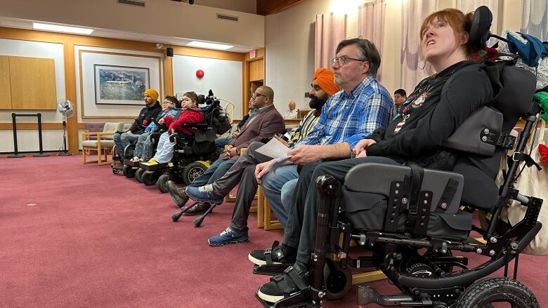 A row of people, some in wheelchairs, sit inside a meeting room.