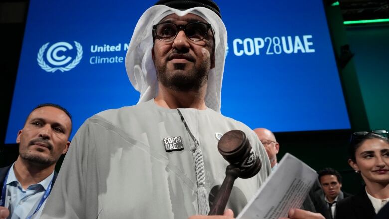 A man with a head scarf and glasses stands in front of a blue screen that reads COP28 UAE.