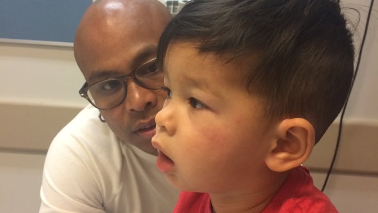 In a hospital room, a man looks concerned at a boy breathing with his mouth open and who has hives on his cheeks.