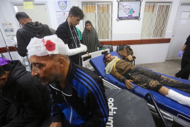 A man with a bloodied bandage around his head sits on a stretcher in a hospital. Another injured man lies on a gurney in the background.