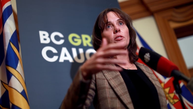 A middle-aged white woman with brown hair cut short wears a brown blazer over a black shirt while speaking at a podium and gesticulating with her hands.