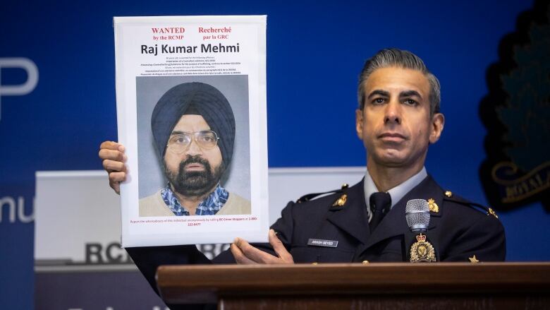 A police officer wearing a navy uniform holds up a wanted posted of Raj Kumar Mehmi, a South Asian man wearing a navy blue turban and glasses.