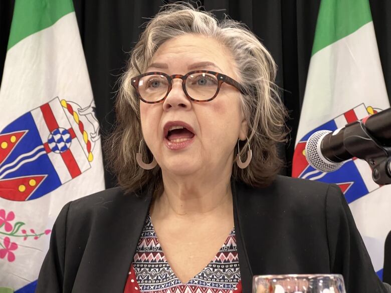 A woman with glasses speaking at a microphone, in front of two flags.