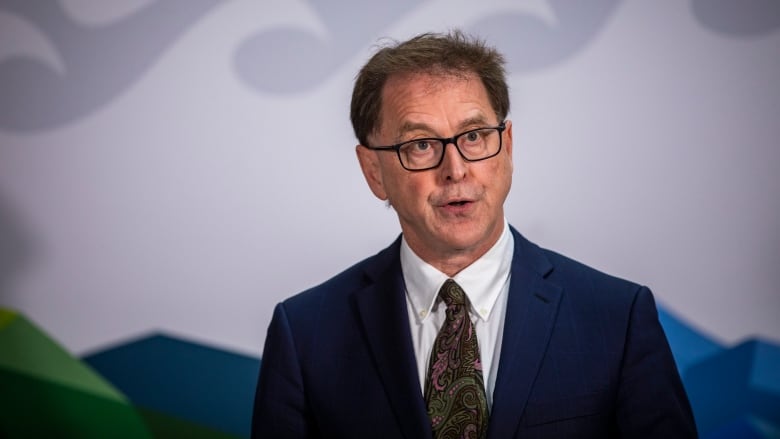 A white man wearing a black tie with fluorescent designs on it speaks while wearing a blue blazer and white shirt.