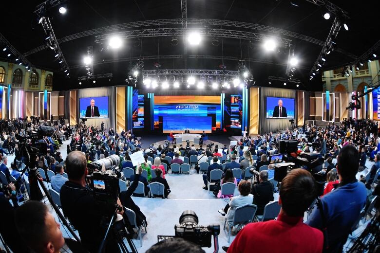 A shot with a fisheye lens shows a large well-lit hall with a large number of people in chairs centred around a distant figure, who is also visible on two large screens to the left and right.